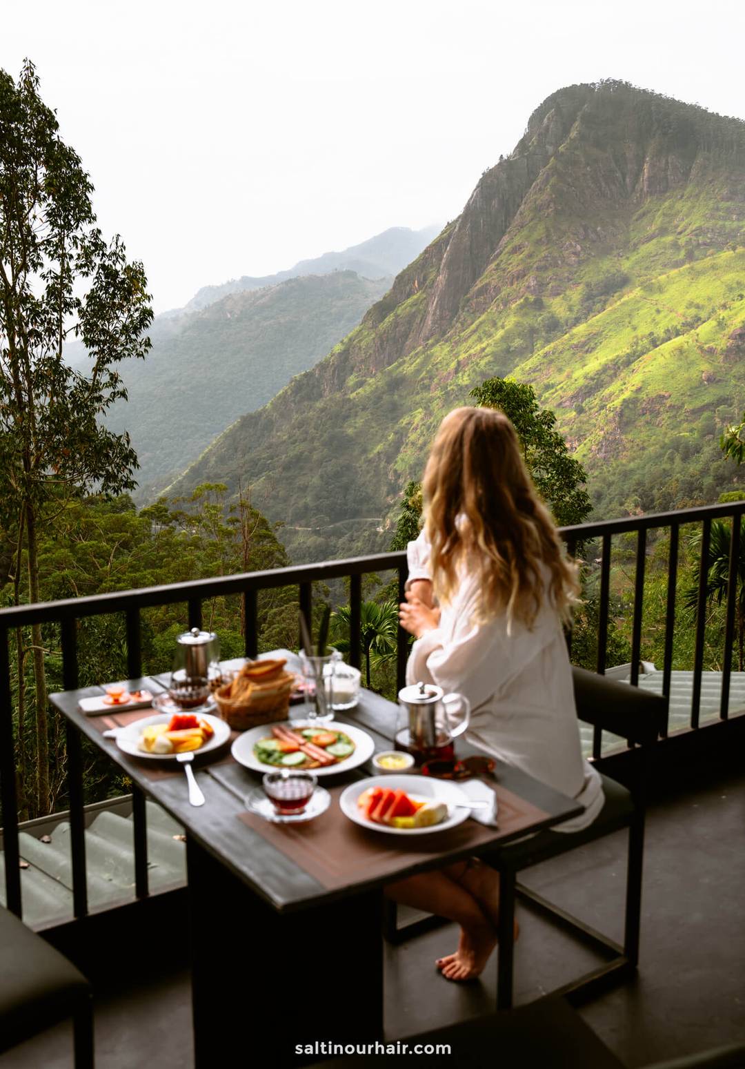 A person sits at a table with breakfast foods, overlooking the lush green mountainous landscape of Ella, Sri Lanka.