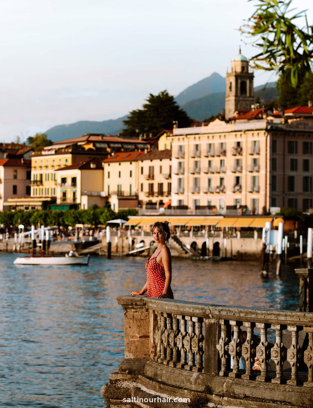 A woman in a red dress stands on a stone balcony by lake como, with colorful buildings and mountains in the background&mdash;an idyllic scene for any italy itinerary 2 weeks adventure.