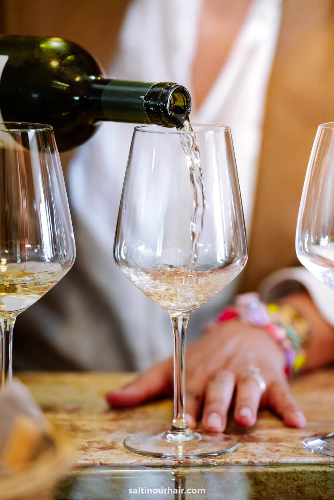 A person pours white wine from a bottle into a clear wine glass on a tabletop, with two empty glasses beside it&mdash;perfect for toasting the start of an unforgettable Italy itinerary 2 weeks adventure.