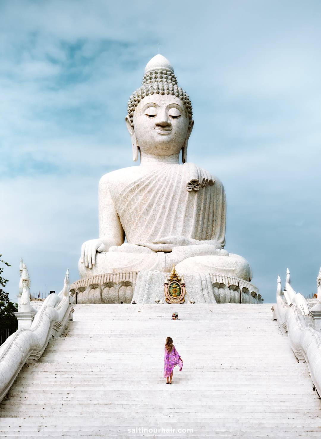 A woman in a pink dress walks up white stairs toward a large seated Buddha statue under a blue sky, capturing the essence of a serene visit to Thailand&mdash;now easier with the new digital arrival card for Thailand.