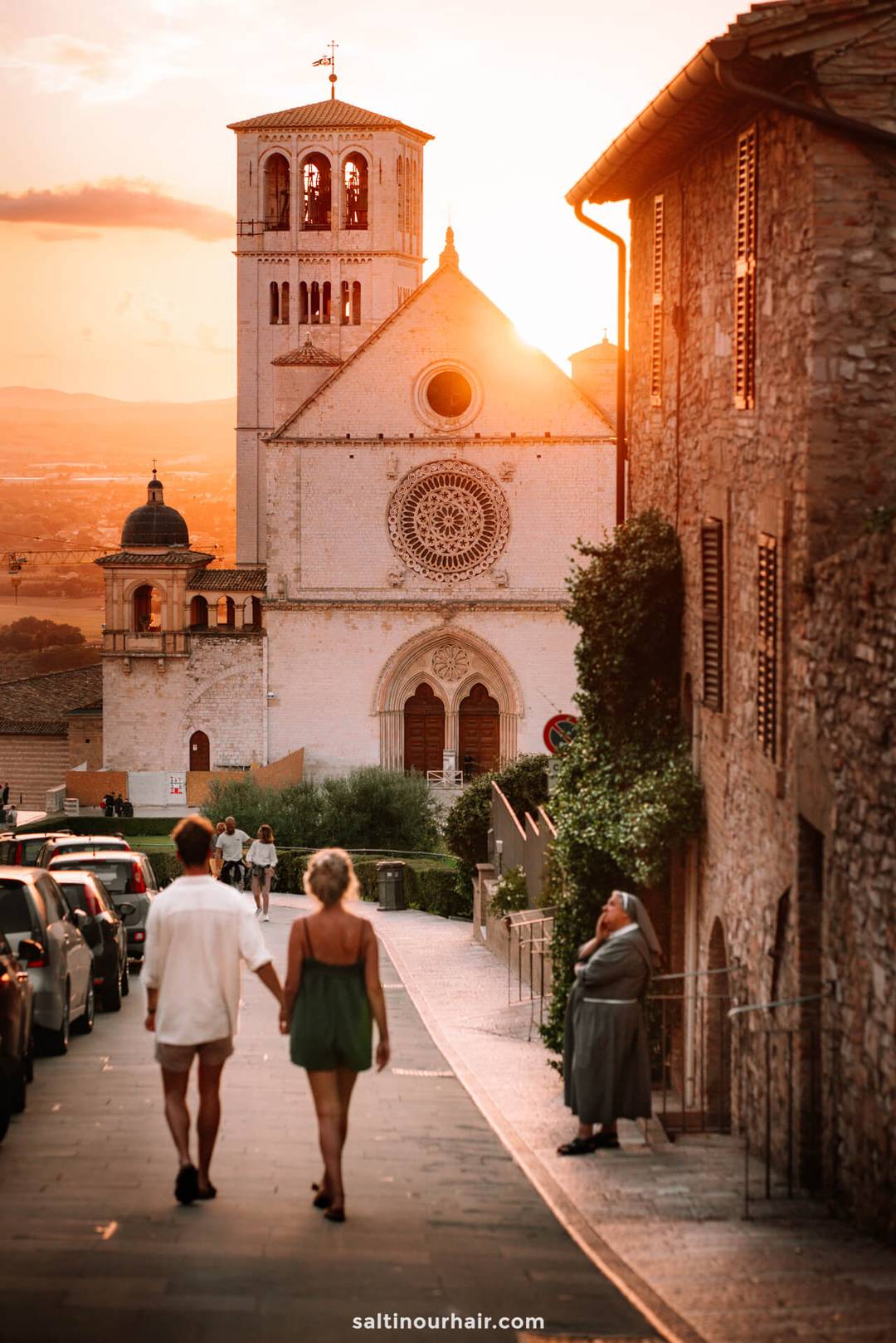 Two people walk hand-in-hand down a street toward a historic church at sunset&mdash;a must-see among things to do on a italy itinerary 2 weeks &mdash;with parked cars and a nun standing by the stone building.