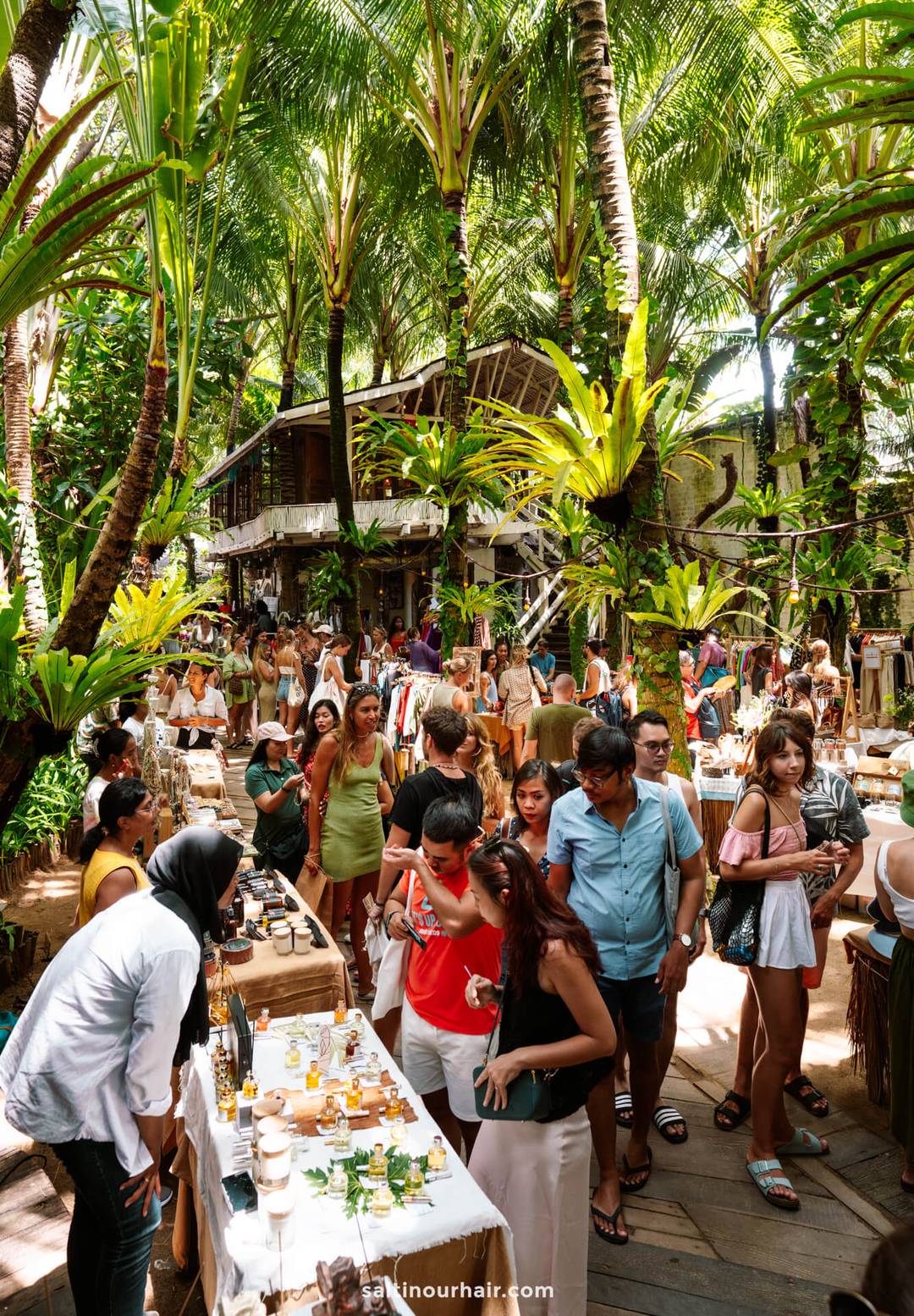 People browse various stalls at the La Brisa Sunday Market in Canggu, Bali, surrounded by tall palm trees and lush greenery, with a rustic wooden building in the background.