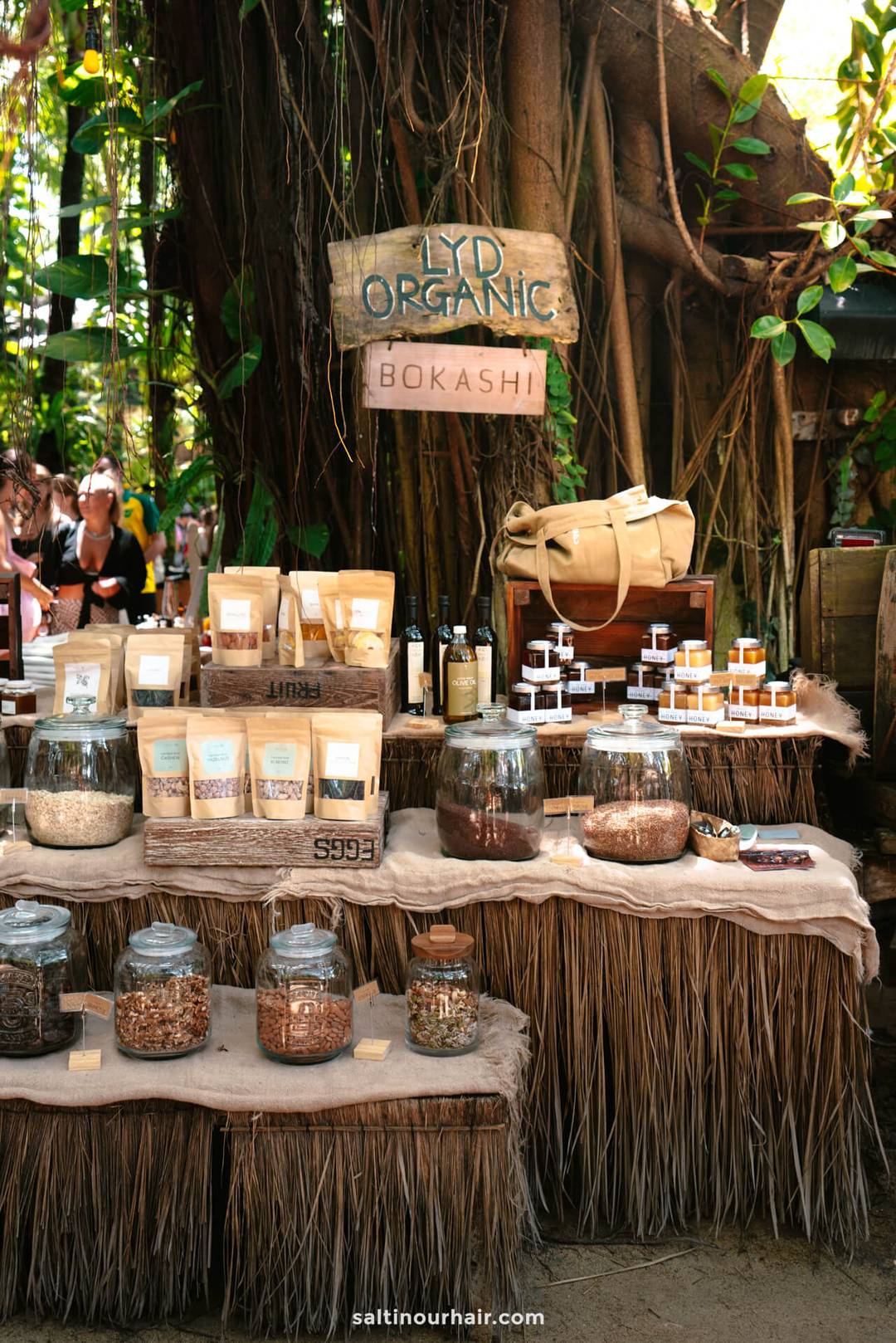 At La Brisa Sunday Market Canggu Bali, a market stall showcases jars, bags, and bottles of organic Bokashi products on tables with natural coverings, greenery all around, and an Organic sign above.