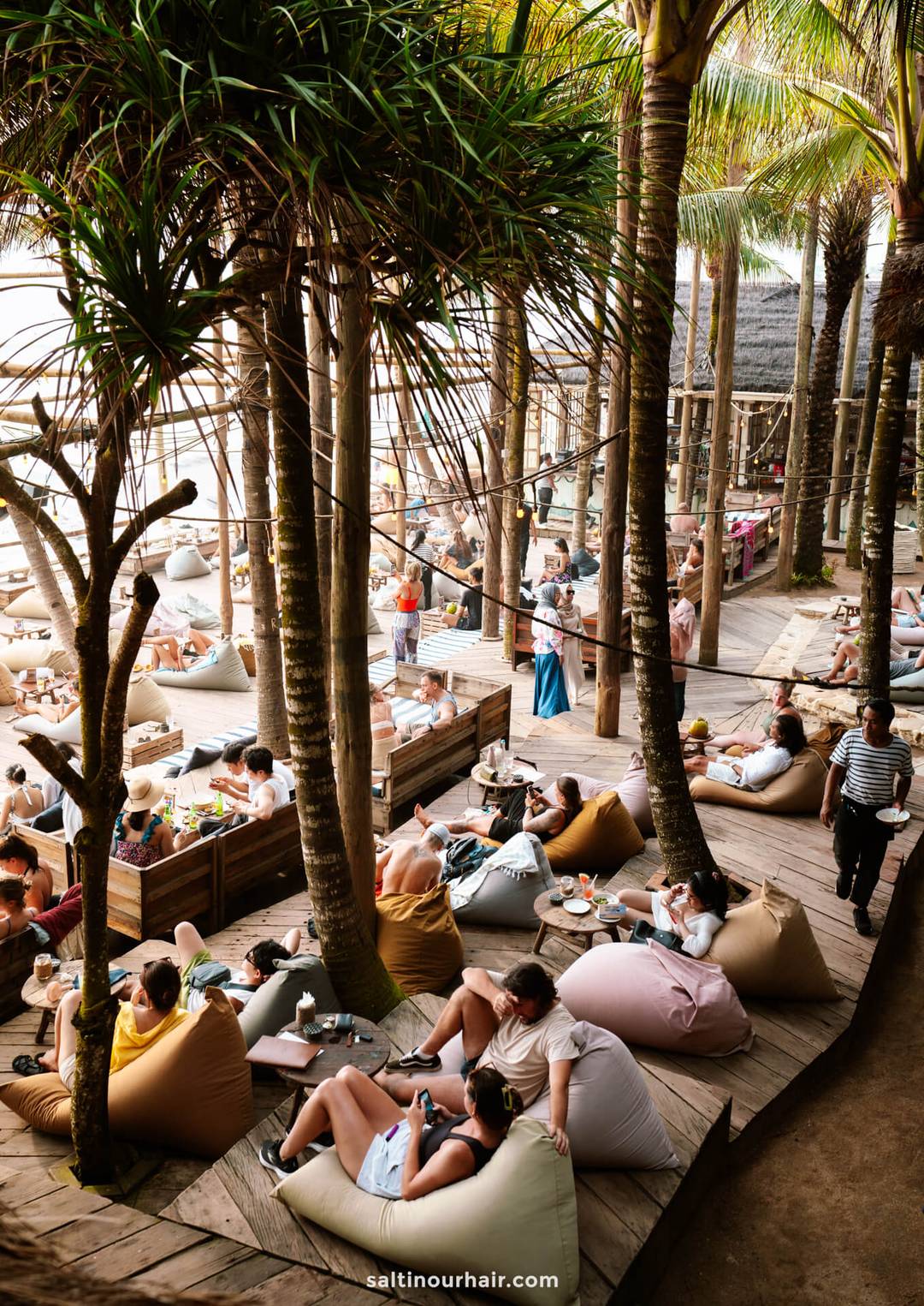 People relax on bean bags and wooden platforms under palm trees at a beachclub in Canggu, Bali, enjoying food and drinks on low tables with a stunning view of the ocean in the background.