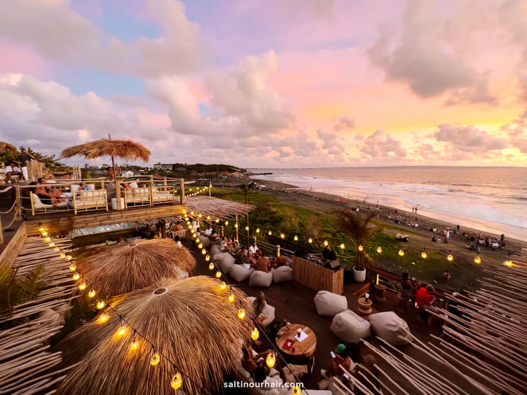 Outdoor beachside restaurant at sunset with people dining, string lights overhead, palm thatched roofs, and ocean waves in the background&mdash;evoking the relaxed vibe of a Canggu Bali beach club.