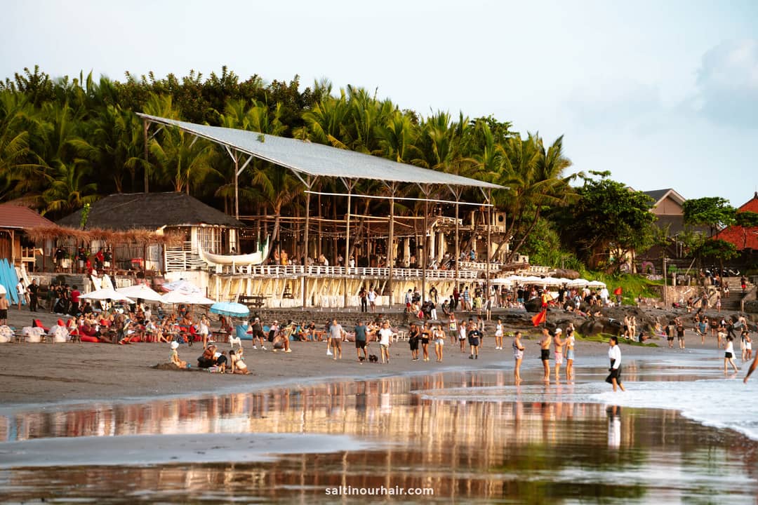 Crowd of people relaxing and walking on a sandy beach near a large, open-air wooden beach bar surrounded by palm trees in vibrant Canggu, Bali. 
