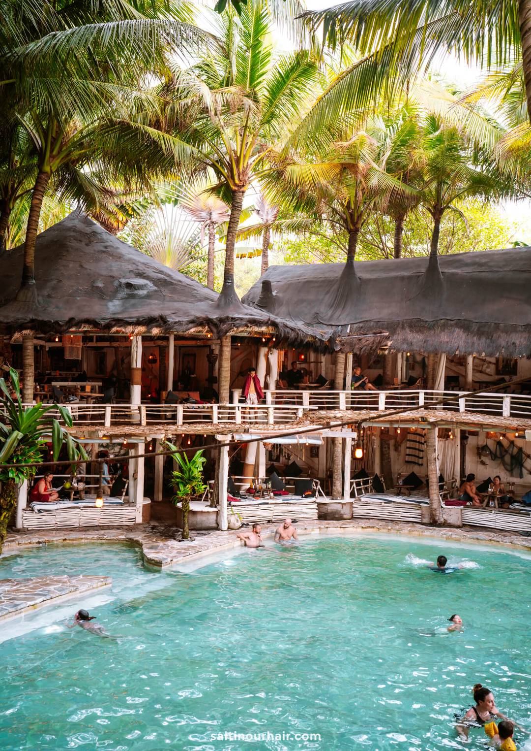 People swim in a turquoise pool surrounded by palm trees and a rustic, two-story building at La Brisa Beach Club, Canggu, Bali, with thatched roofs and wooden railings.