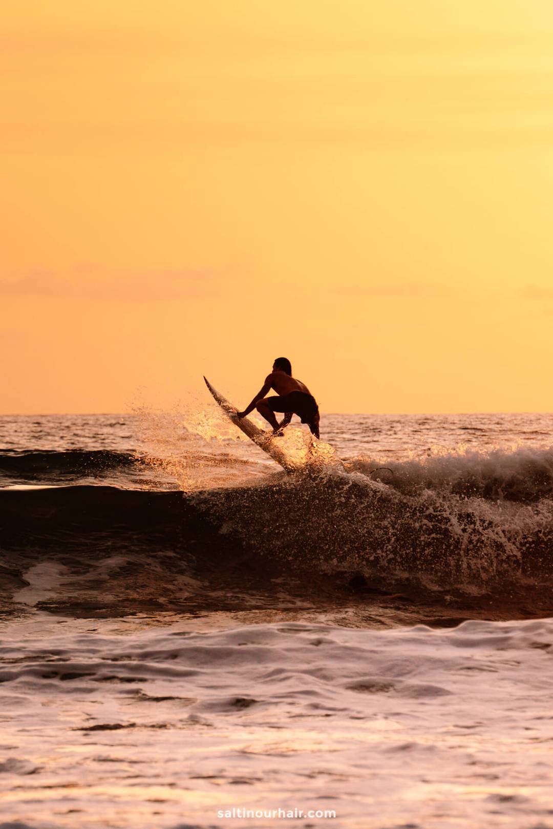 A surfer rides a wave at sunset in Canggu, Bali, silhouetted against the orange sunset sky, with ocean spray surrounding the surfboard.