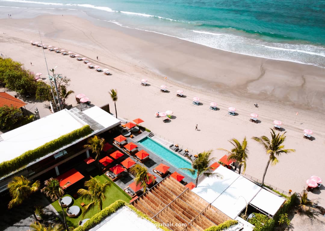 Aerial view of Seminyak Beach in Bali, featuring rows of pink umbrellas, a rectangular pool, green lawn, palm trees, and nearby buildings along the scenic shoreline near Canggu.