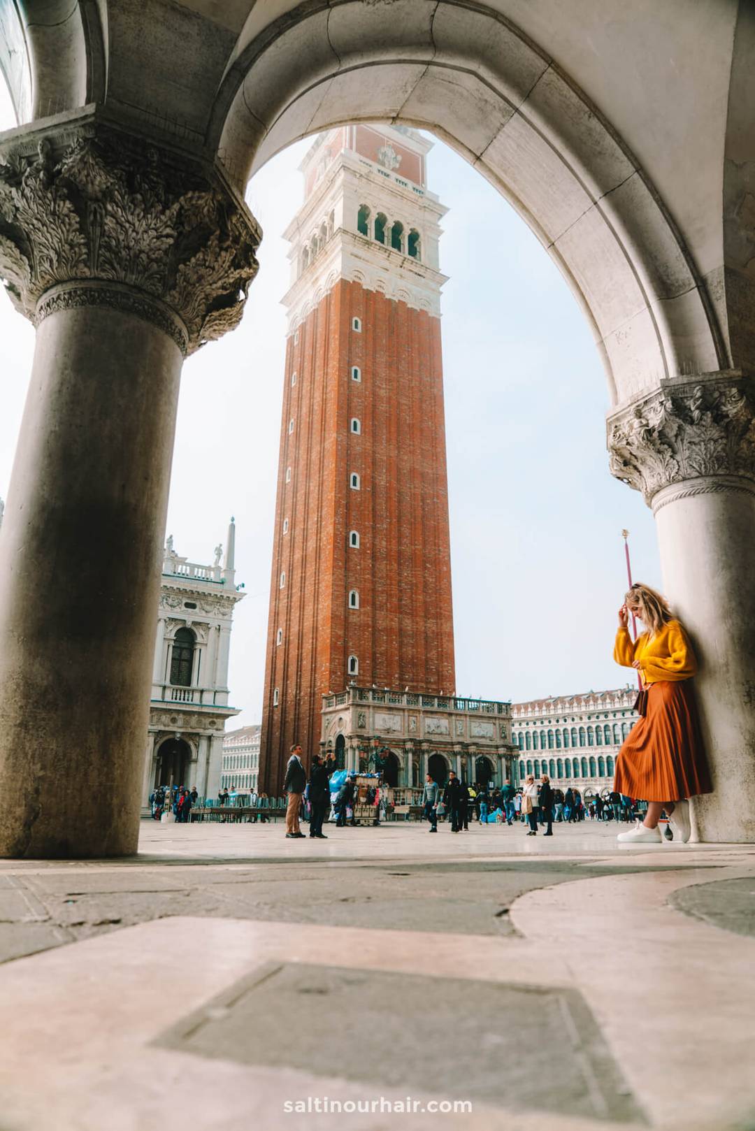 A tall red brick bell tower stands in a busy square, framed by stone arches; a woman in an orange skirt and yellow top walks in the foreground&mdash;perfect inspiration for your Italy 7 day itinerary.