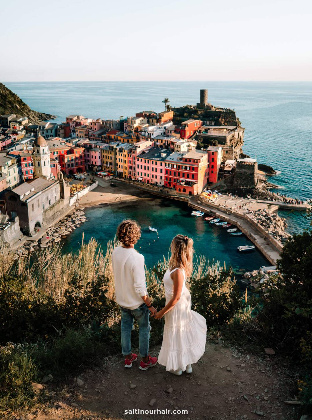 A couple stands on a hill overlooking the colorful coastal village of Vernazza, a highlight on any Italy 7 day itinerary, with the sparkling sea and historic buildings visible below.