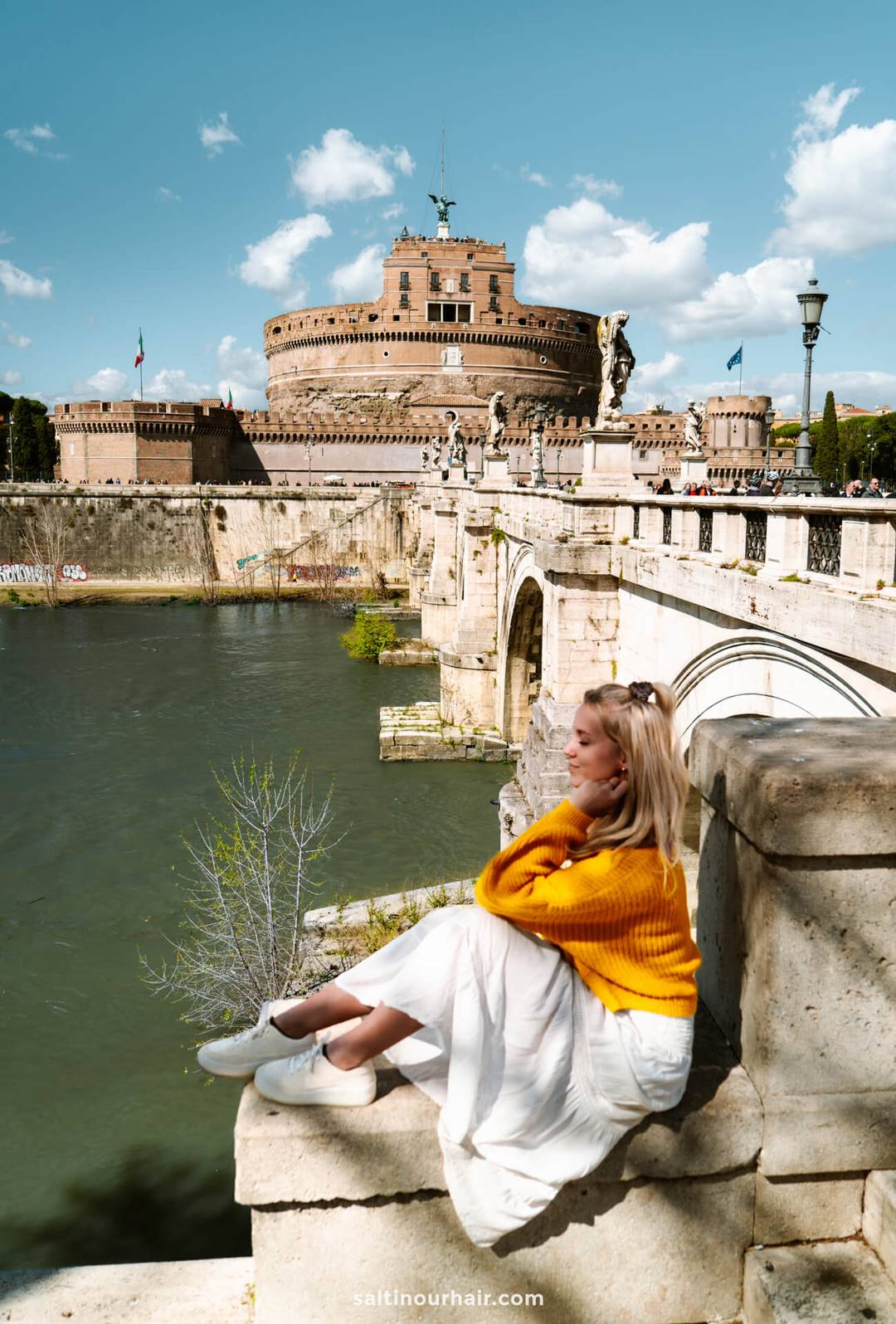 A woman in a yellow sweater and white skirt sits by the Tiber River with Castel SantAngelo and a stone bridge in the background&mdash;perfect inspiration for your Italy 7 day itinerary under a partly cloudy sky.