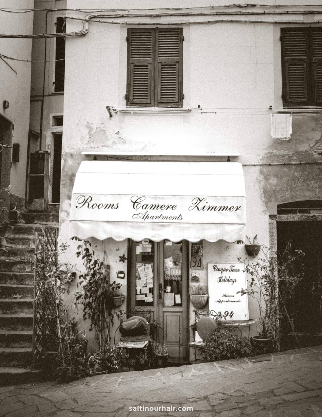 Black and white photo of a building facade in Italy, featuring a striped awning reading Rooms Camere Zimmer Apartments above a door, surrounded by plants&mdash;a scene you might spot on your Italy 7 day itinerary.