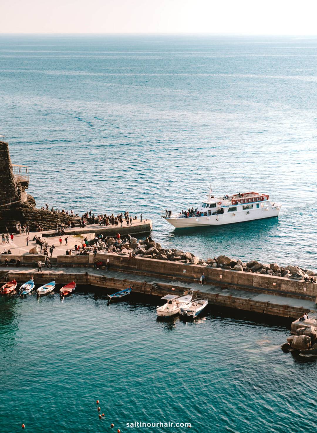 A white ferry approaches a stone pier crowded with people, with several small boats docked in the foreground and calm blue sea in the background&mdash;a classic scene on an Italy 7 day itinerary.