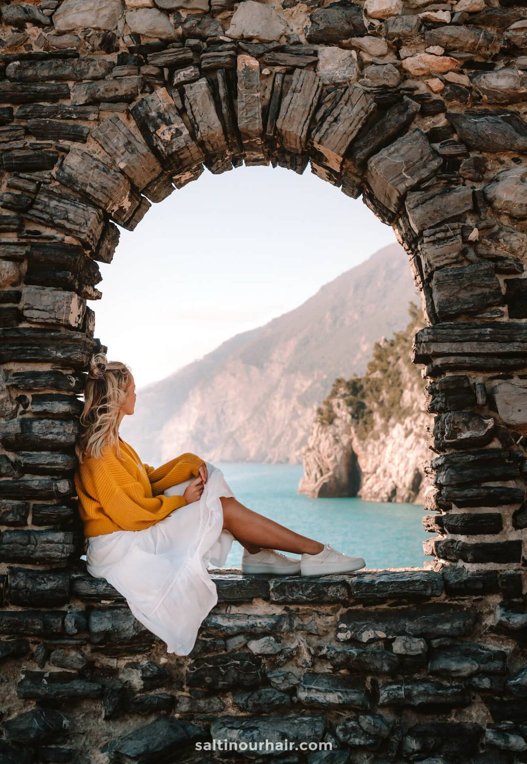 Woman in a yellow sweater and white skirt sits on a stone windowsill, gazing at the sea and mountains through an arched opening&mdash;capturing the serene beauty perfect for any Italy 7 day itinerary.