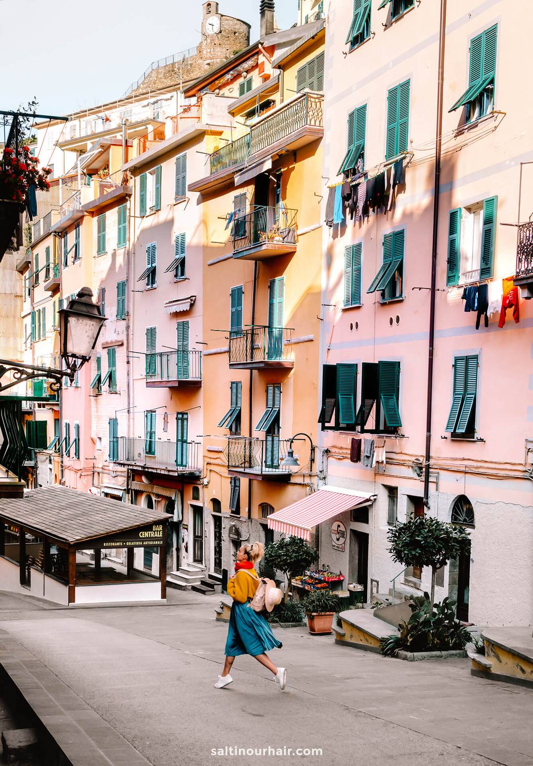 A woman walks on a street in Italy, lined with colorful buildings featuring green shutters and balconies, some with laundry hanging outside&mdash;a picturesque scene perfect for an Italy 7 day itinerary.