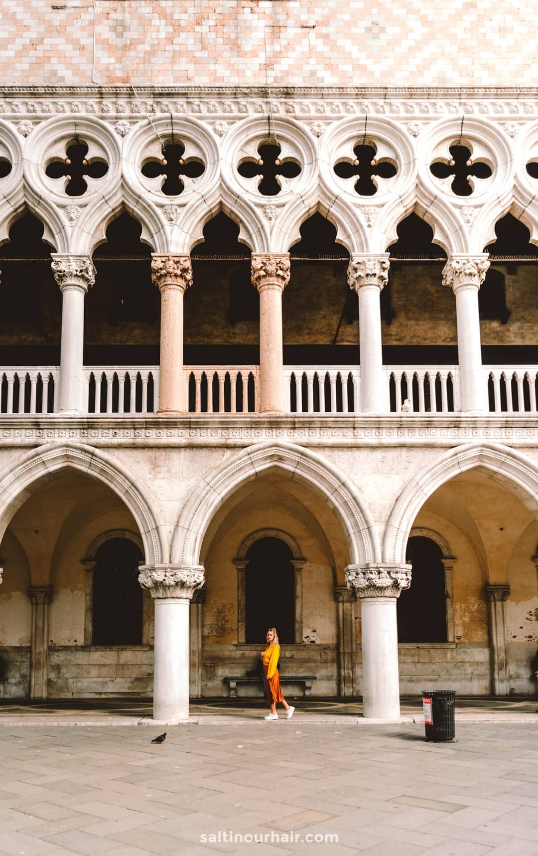 A woman in a yellow dress walks in front of an ornate, arched stone building with columns and a patterned facade&mdash;perfect inspiration for your Italy 7 day itinerary.