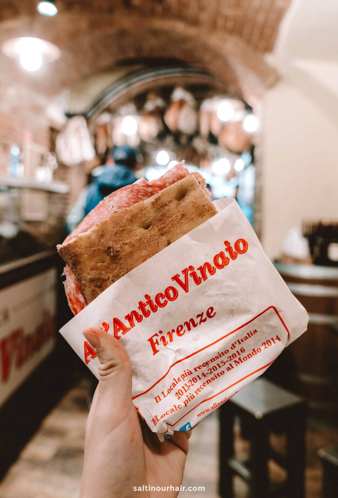 A hand holds a sandwich wrapped in paper labeled All&rsquo;Antico Vinaio Firenze inside a deli with exposed brick walls&mdash;an iconic stop on any Italy 7 day itinerary.