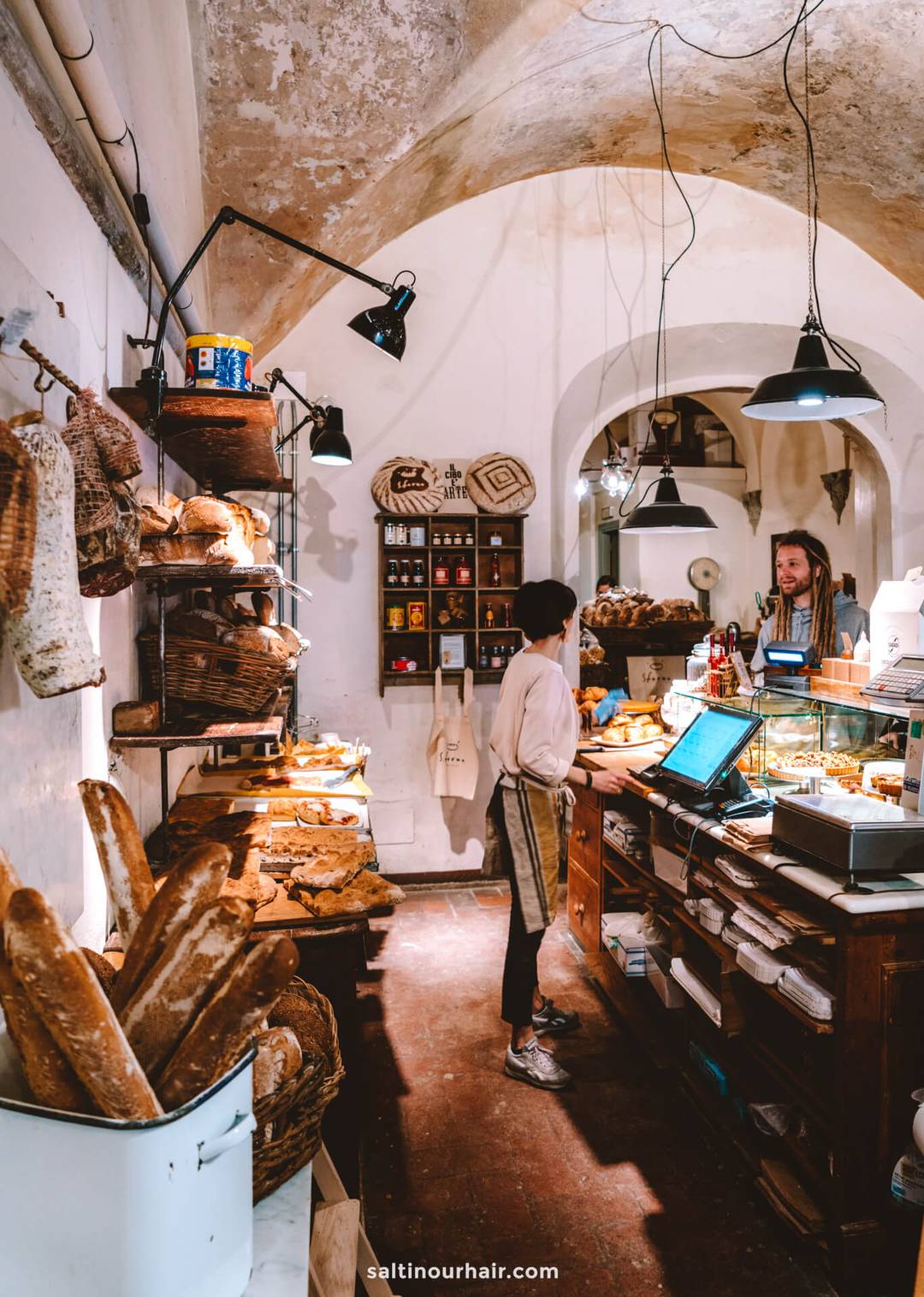 A woman stands at the counter of a rustic bakery in Italy, surrounded by shelves of bread, baskets, and hanging cured meats&mdash;an authentic stop on any Italy 7 day itinerary&mdash;as another person serves her.