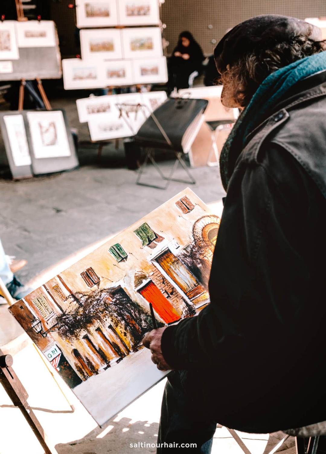 An artist sits outdoors painting a colorful street scene on a canvas, with completed artworks displayed on easels behind them&mdash;capturing the vibrant charm you