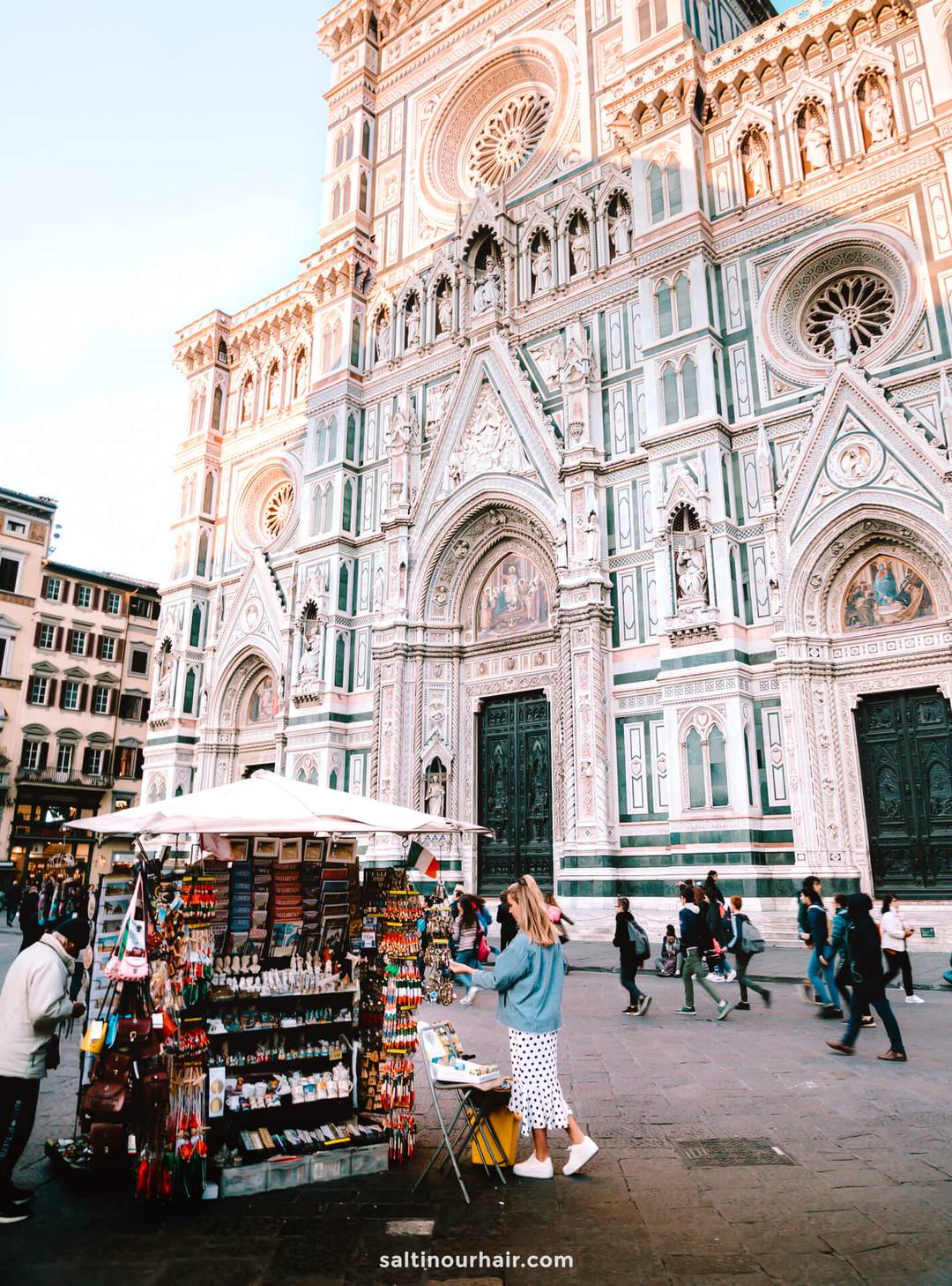 A woman stands at a souvenir stall in front of the ornate facade of Florence Cathedral, a must-see stop on any Italy 7 day itinerary, while people walk by in the bustling square.