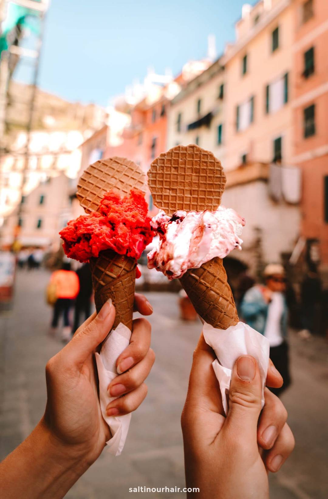 Two hands holding waffle cones with scoops of red and white gelato, topped with waffle cookies, on a charming street in Italy&mdash;perfect inspiration for your Italy 7 day itinerary.