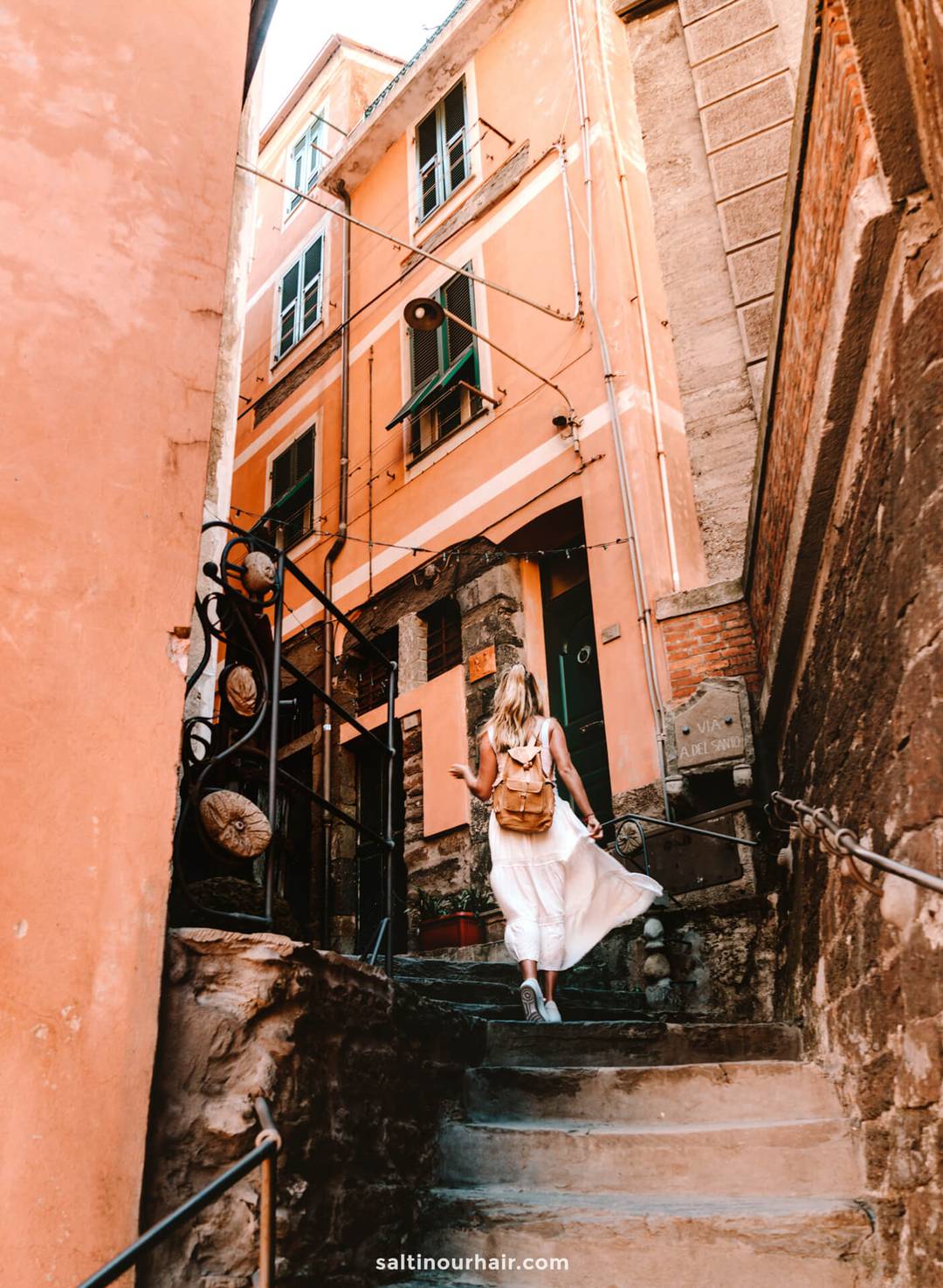 A woman with a backpack and white dress walks up narrow stone steps between orange buildings in a European alleyway, capturing the spirit of discovery found on an Italy 7 day itinerary.