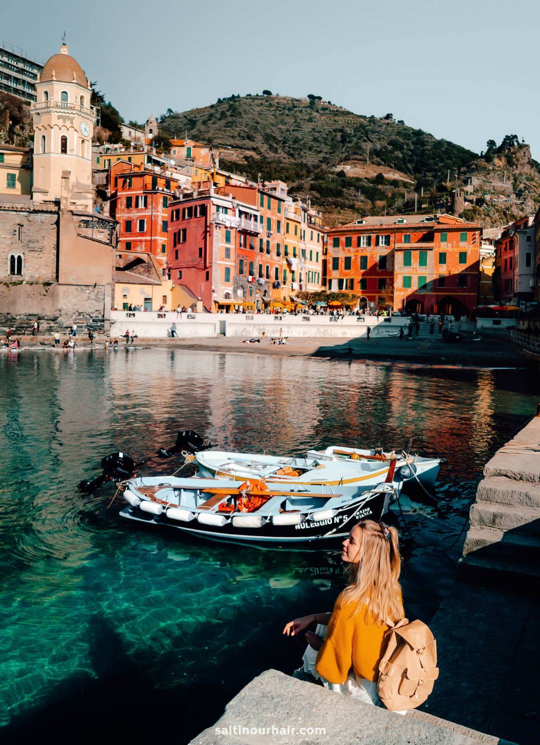 A woman with a backpack sits by clear water facing colorful buildings and boats docked in a small harbor, hills behind her&mdash;a perfect scene from an Italy 7 day itinerary.