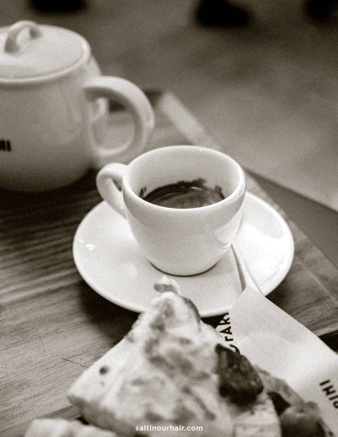 A white espresso cup and saucer sit on a wooden table next to a teapot and a plate of food in this black-and-white photo, evoking the classic charm of an Italy 7 day itinerary.