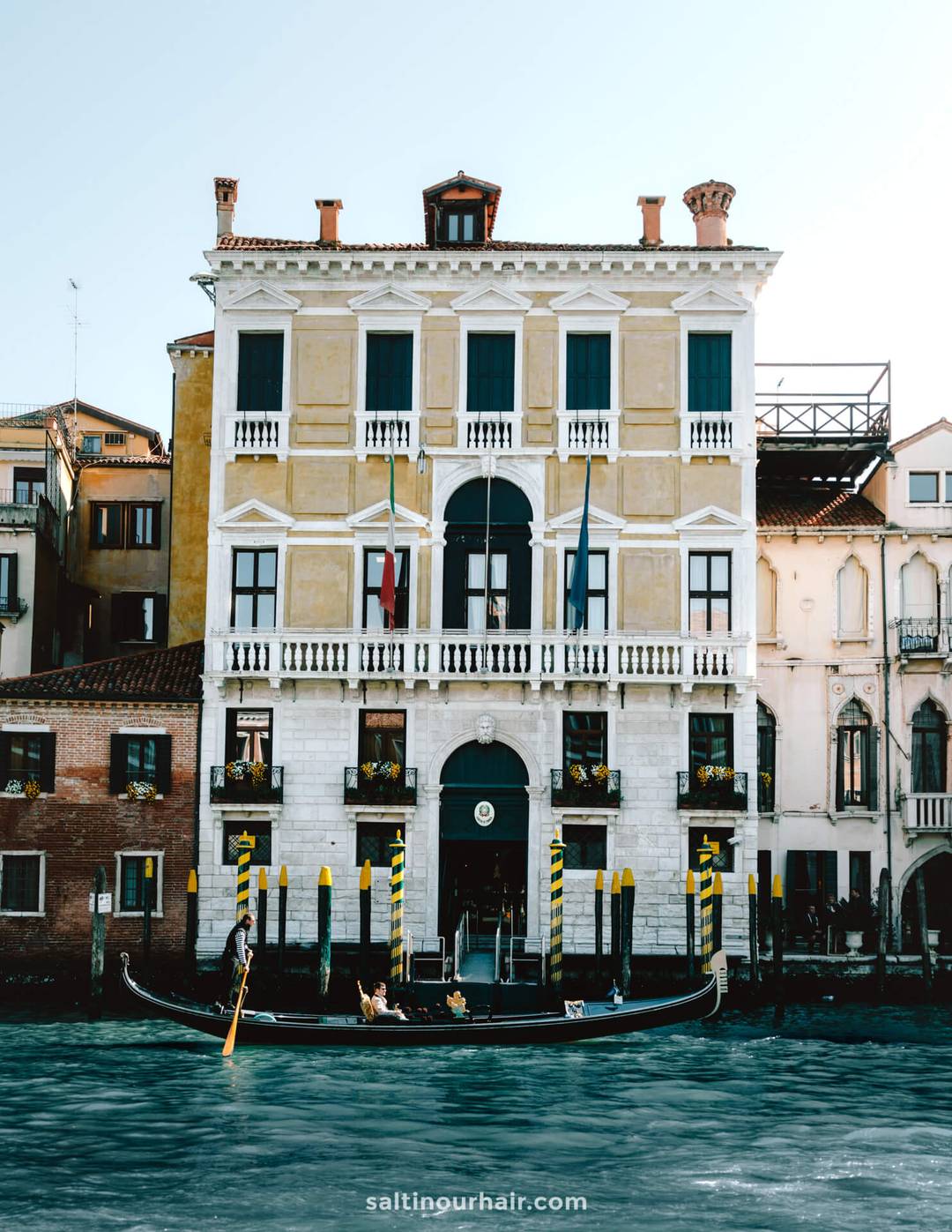 A gondola with passengers floats on a canal in front of a historic yellow building with arched windows and flags&mdash;an iconic scene to include in your Italy 7 day itinerary while exploring the charms of Venice.