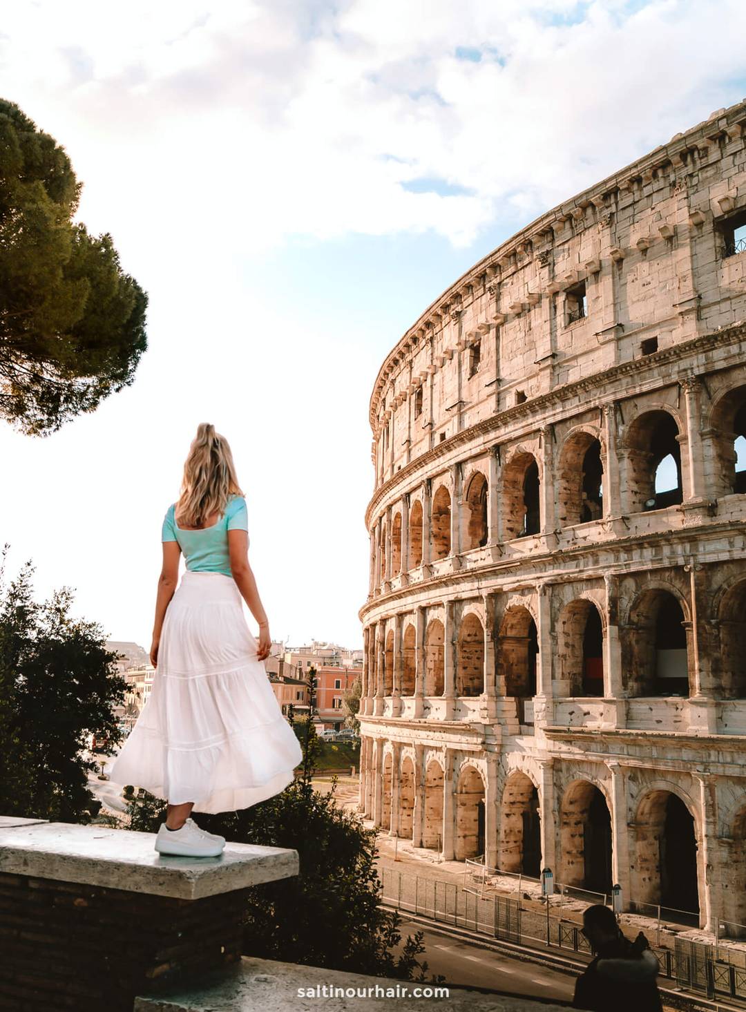 A woman in a blue top and white skirt stands on a ledge, gazing at the Colosseum in Rome under a partly cloudy sky&mdash;an iconic moment on any italy itinerary 2 weeks