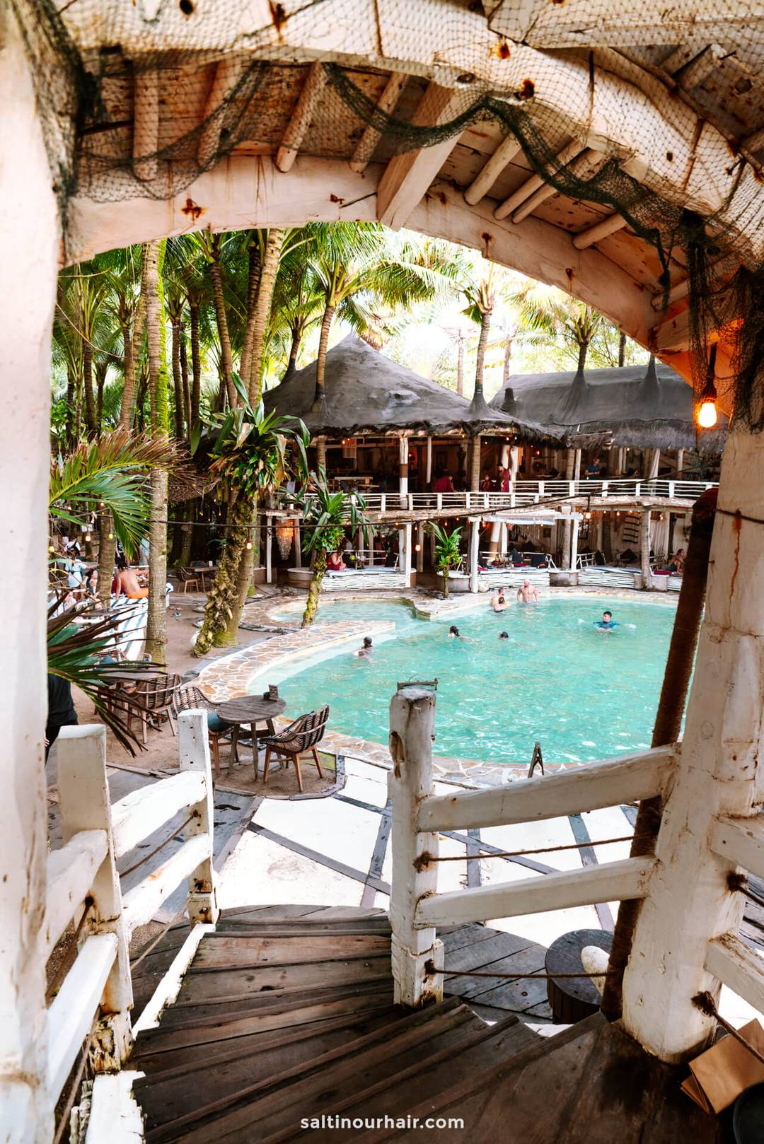 View from a wooden staircase overlooking the tropical resort pool at La Brisa Canggu Bali, surrounded by palm trees, lounge chairs, and people swimming.