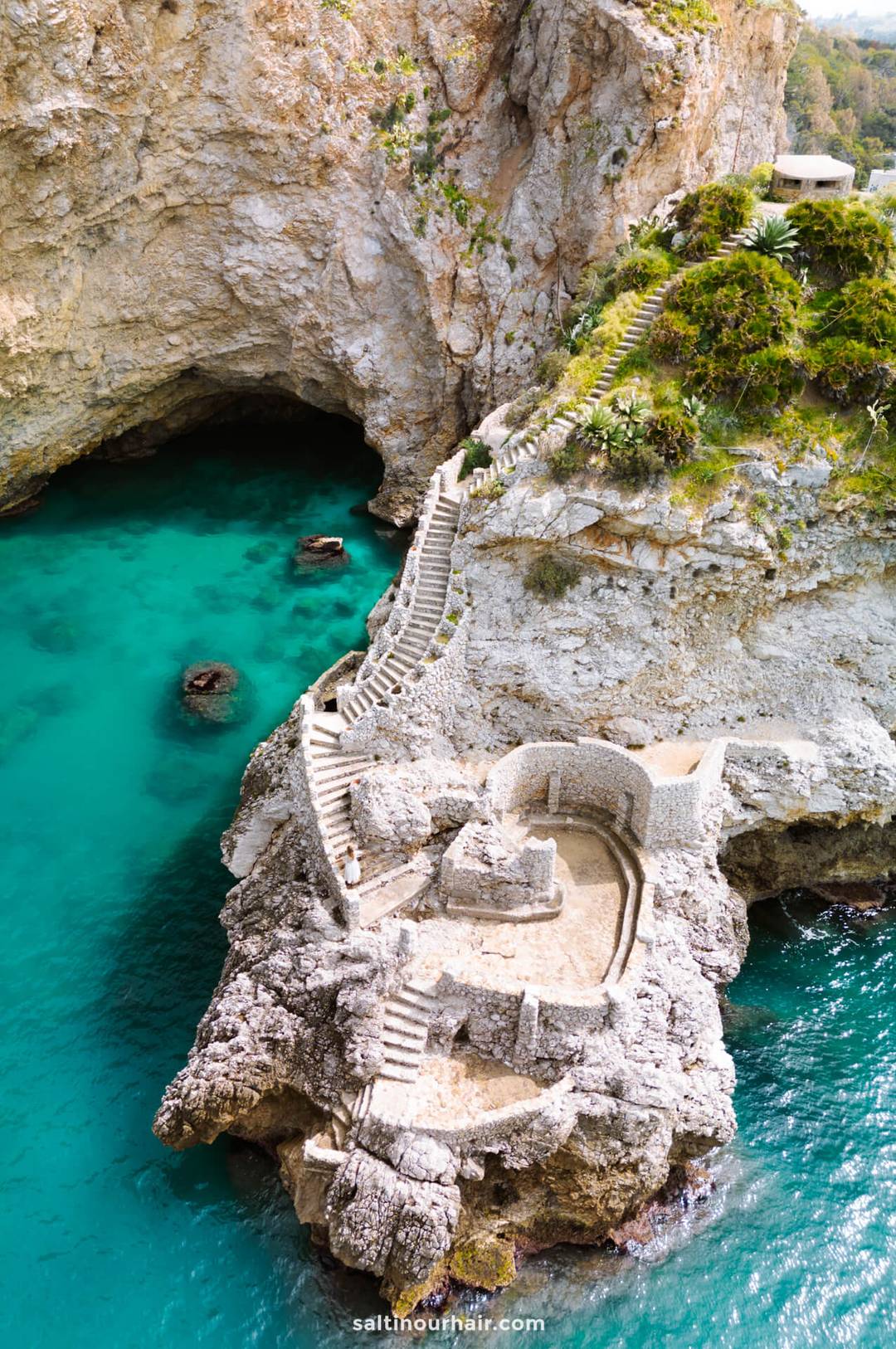 A stone staircase and pathways wind along rocky cliffs above turquoise water at the Cueva de Los Tres Ojos, evoking the adventure found in places like Bunker de San Cataldo, one of the top things to do near Trapani, Sicily.