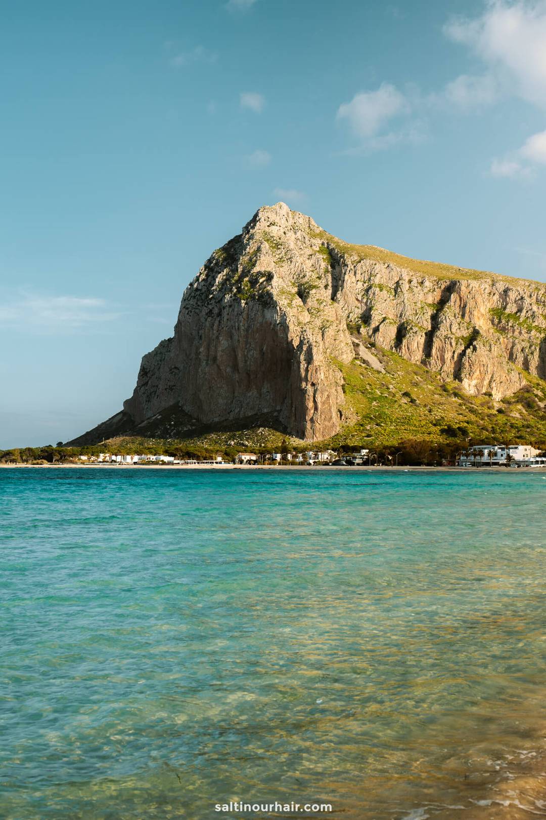 Coastal scene at San Vito Lo Capo, Sicily, Italy, with clear blue water in the foreground and a large rocky mountain rising near the shoreline under a partly cloudy sky.