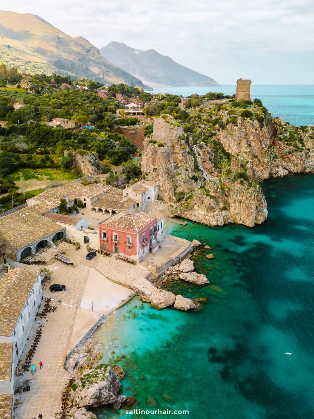 Aerial view of Scopello, Sicily&mdash;a coastal village with stone buildings, clear turquoise water, rocky cliffs, and a tower on a hill overlooking the sea.