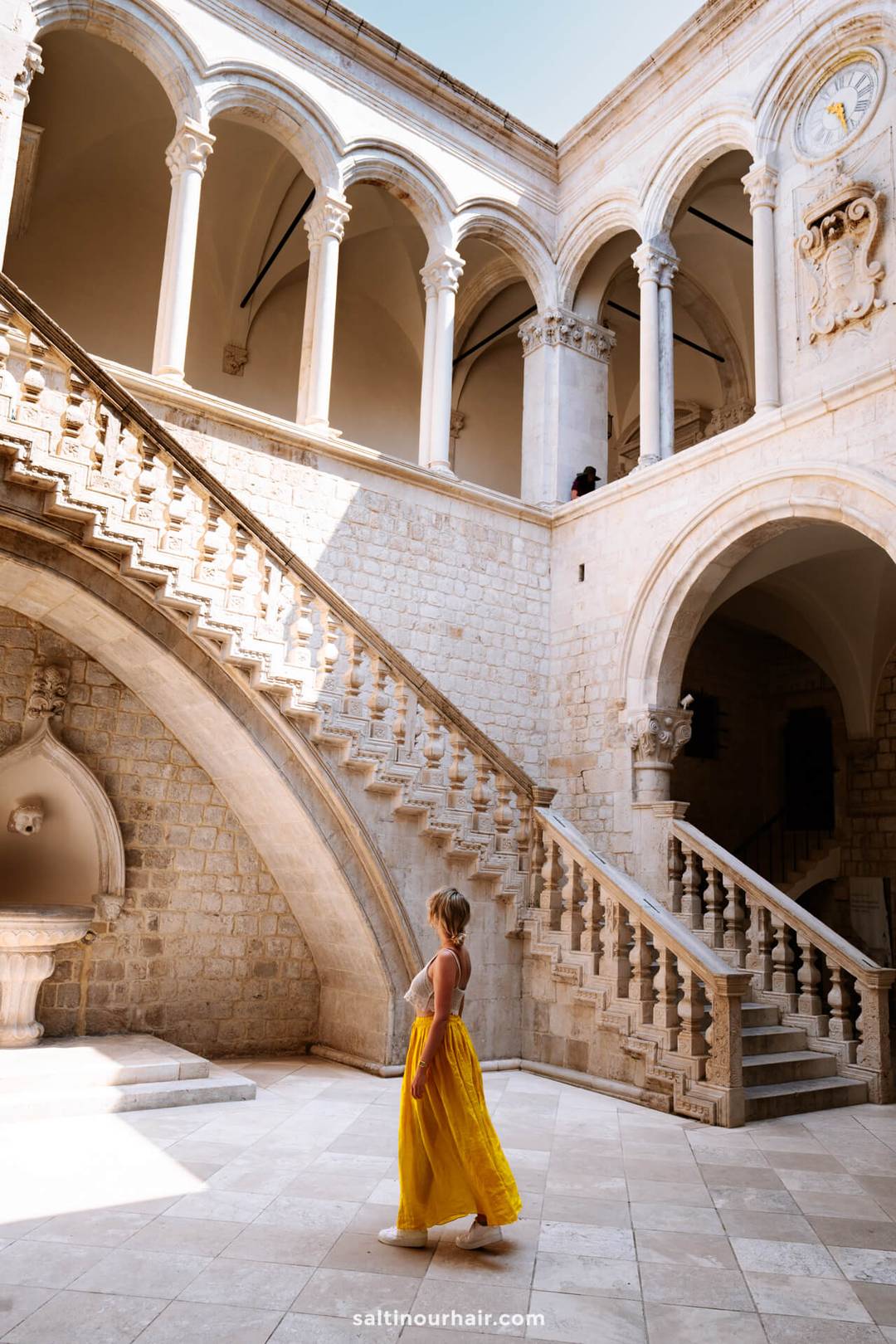 A woman in a yellow dress stands at the base of a grand stone staircase in a historic, sunlit courtyard with arches and columns, capturing the charm of Dubrovnik, Croatia on an unforgettable road trip.
