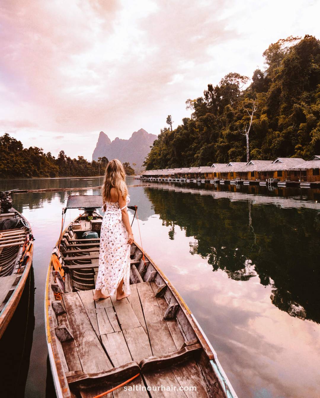 A woman in a white floral dress stands on a wooden boat on a calm lake in Thailand, surrounded by lush trees and floating huts under a cloudy sky, savoring the serenity just after completing her new digital arrival card for Thailand.