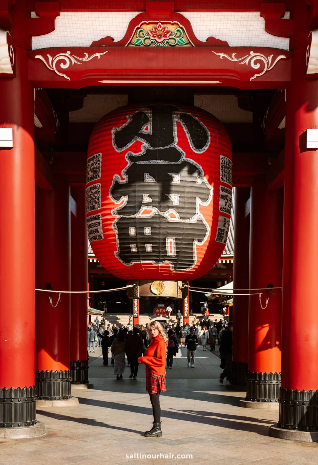 A woman stands in front of a large red lantern at the entrance of Sensō-ji, Tokyo, Japan, surrounded by red columns and visitors in the background.