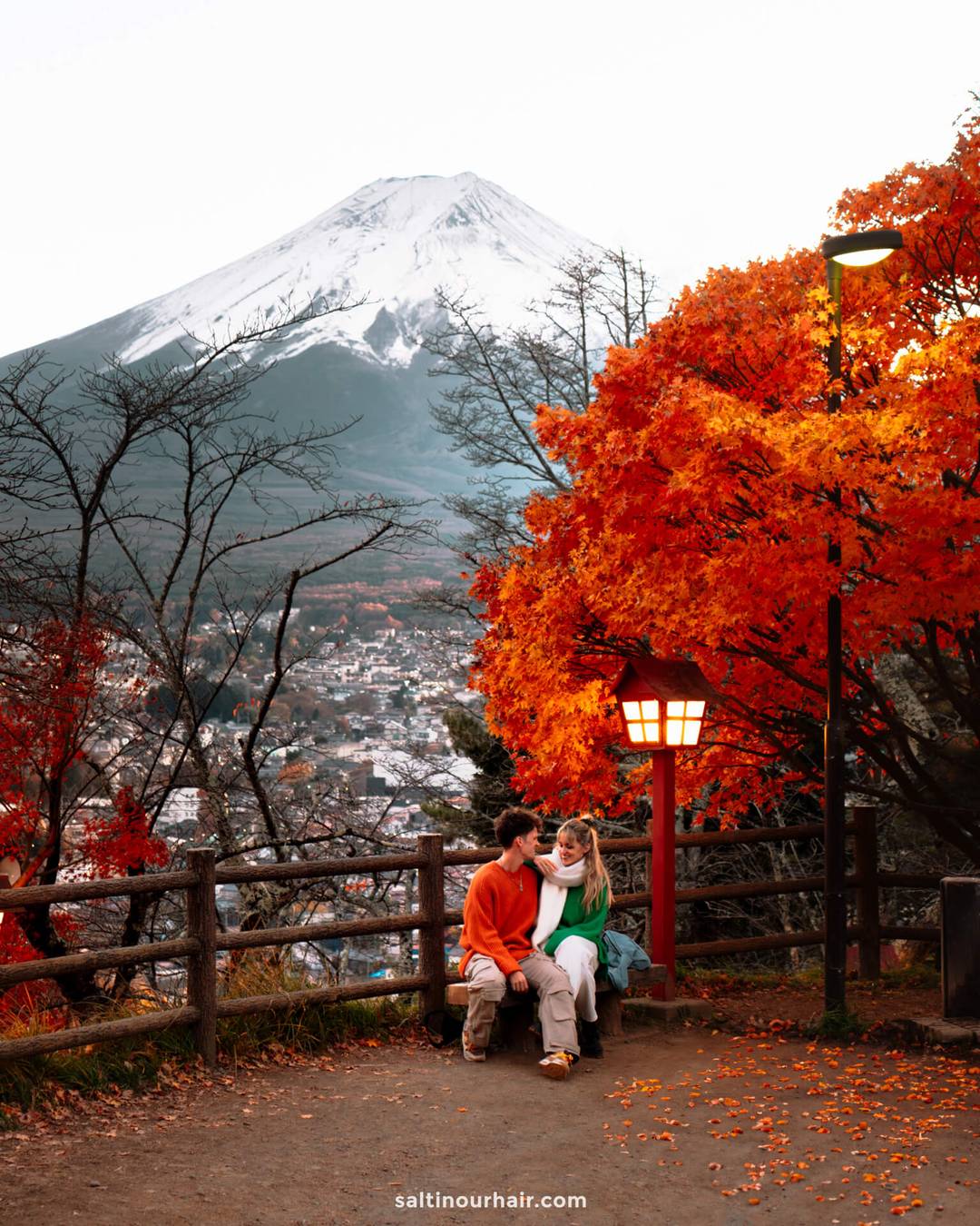 A couple sits on a bench under vibrant red autumn trees&mdash;one of the best places in Japan to see autumn foliage&mdash;with mount fuji in the background and a lit lantern nearby.