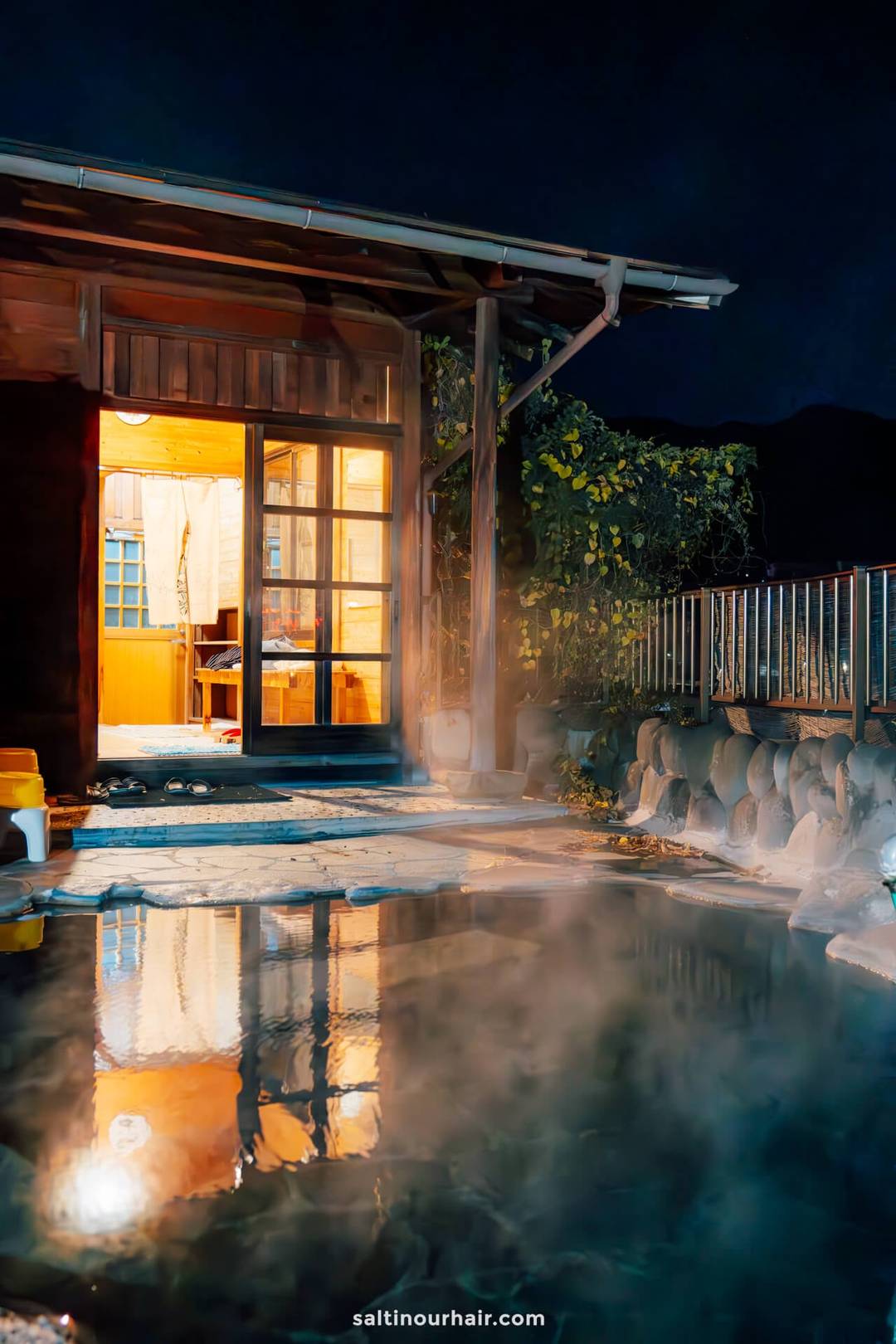 Outdoor Japanese onsen bath with steam rising, adjacent to a wooden building glowing with warm interior lighting at night&mdash; perfect after a day exploring Japan in autumn.