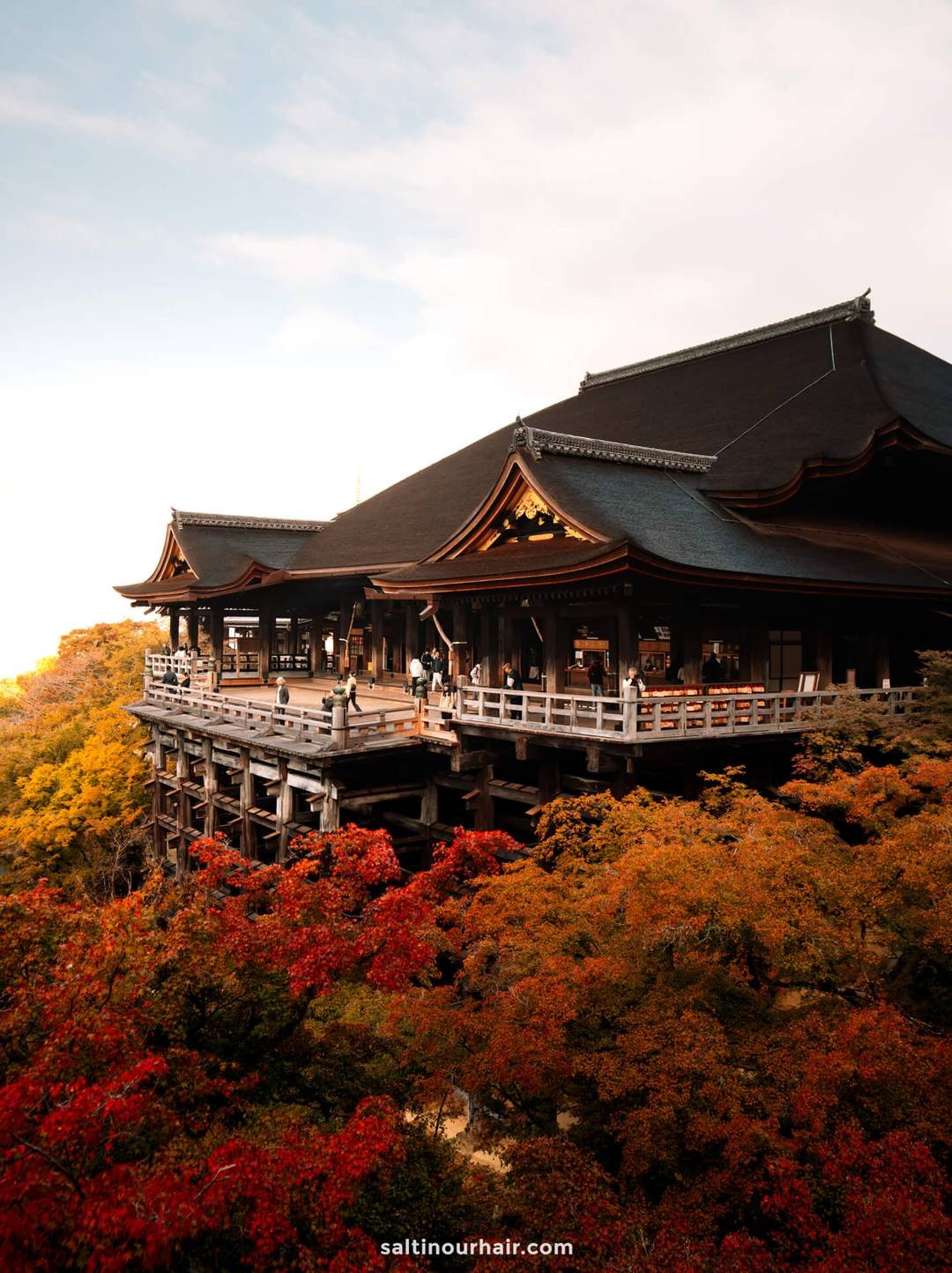 The large wooden Kiyomizu-dera Temple in Japan, with its tiered roofs, overlooks vibrant autumn foliage under a partly cloudy sky. Several people are visible on the terrace.