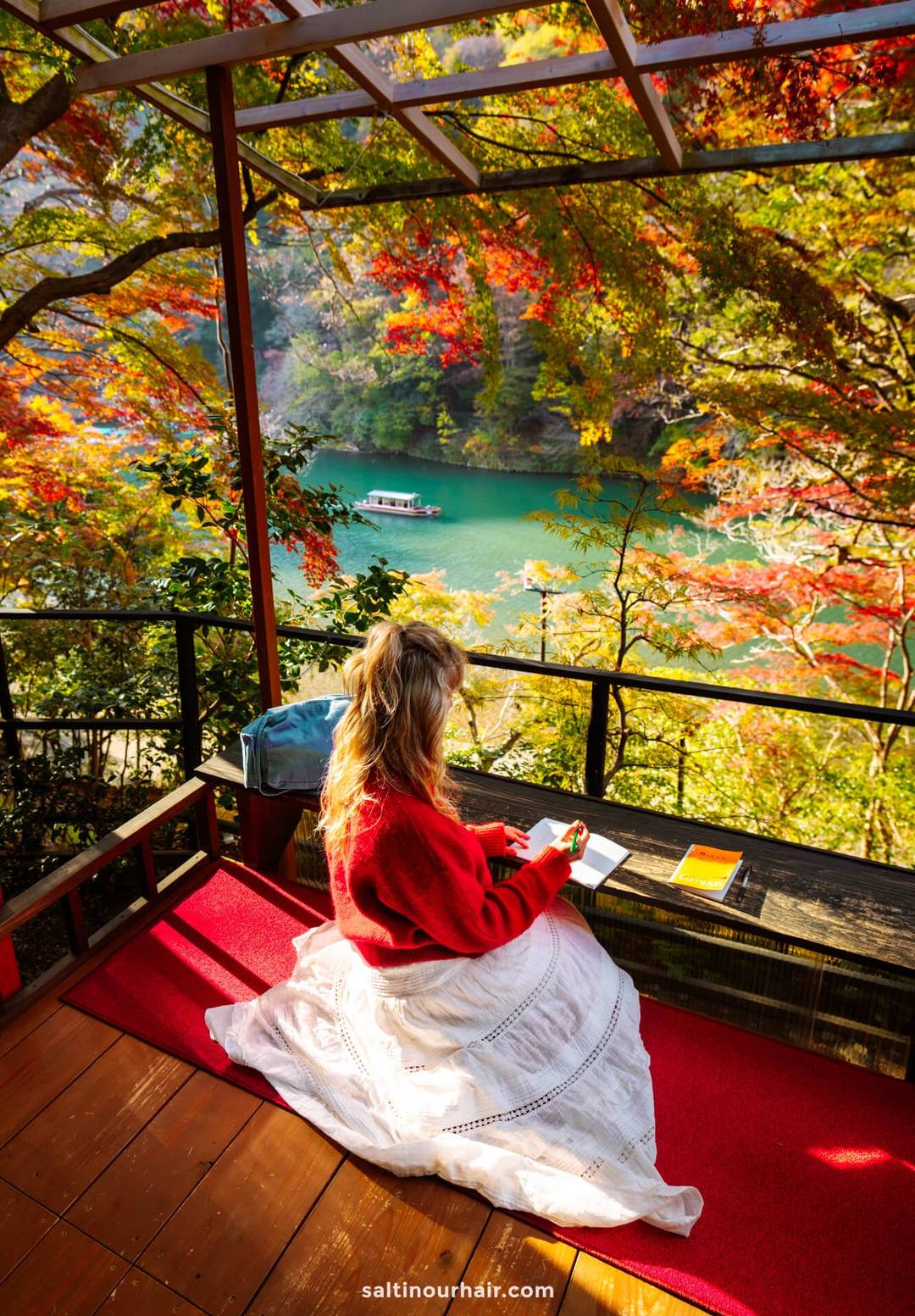 A woman in a red sweater and white skirt sits on a terrace at Yusai-Tei Gallery in Kyoto, Japan, overlooking a river with autumn foliage and a passing boat in the background.