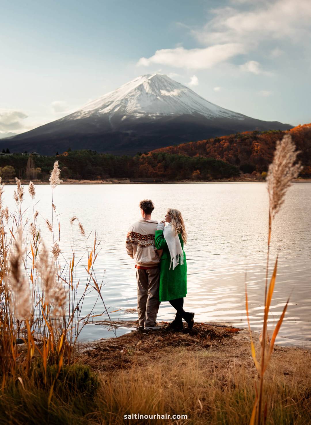 A couple stands by a lakeshore facing snow-capped Mount Fuji in Japan, with tall grasses in the foreground and autumn trees in the background.