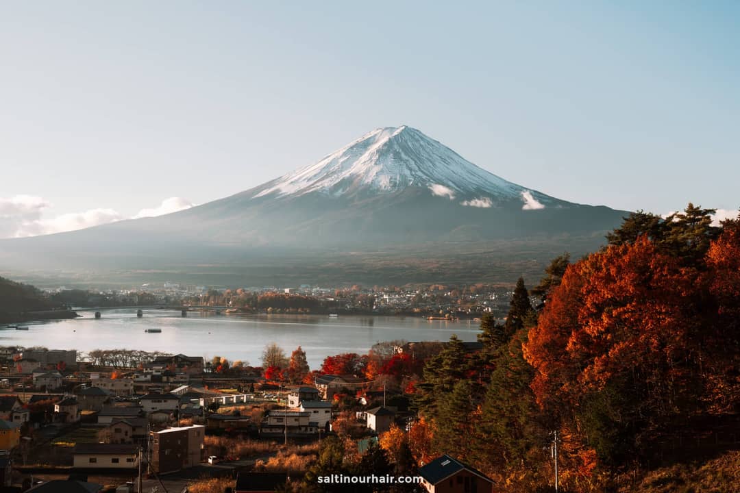 Snow-capped Mount Fuji rises above a tranquil Japanese town and lake, with vibrant autumn trees in the foreground under a clear sky.