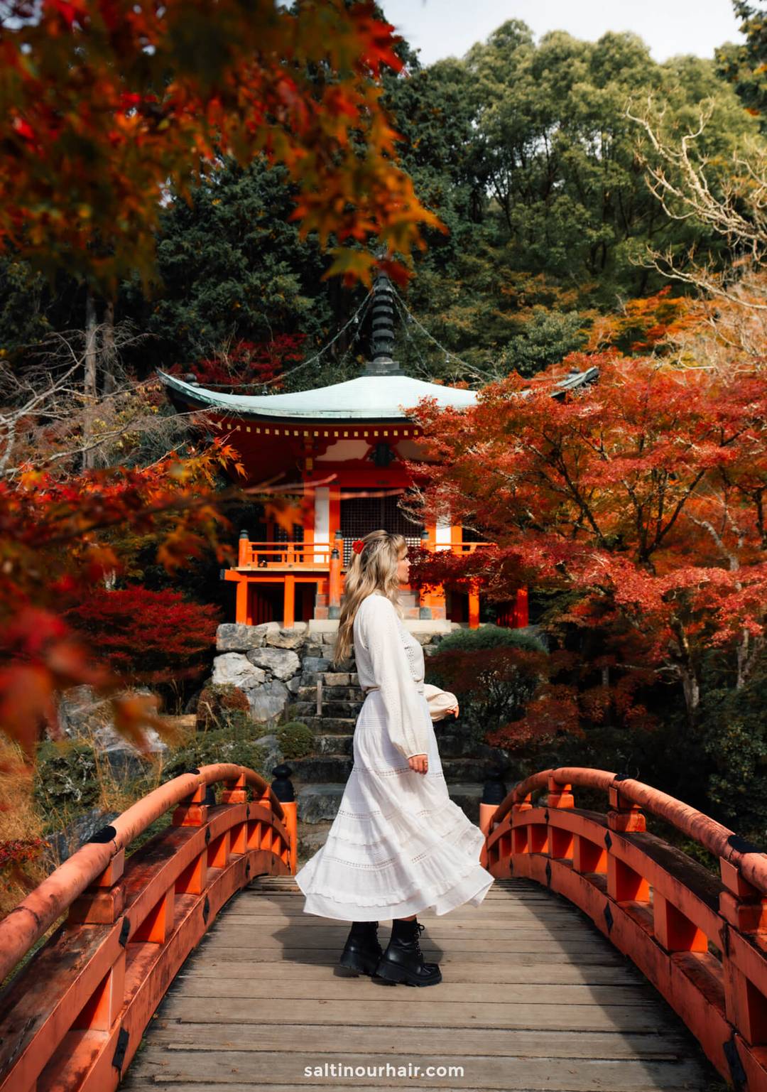 A woman in a white dress stands on a red wooden bridge, facing a traditional Japanese pavilion at Daigo-ji Temple, surrounded by colorful autumn trees during autumn in Japan.