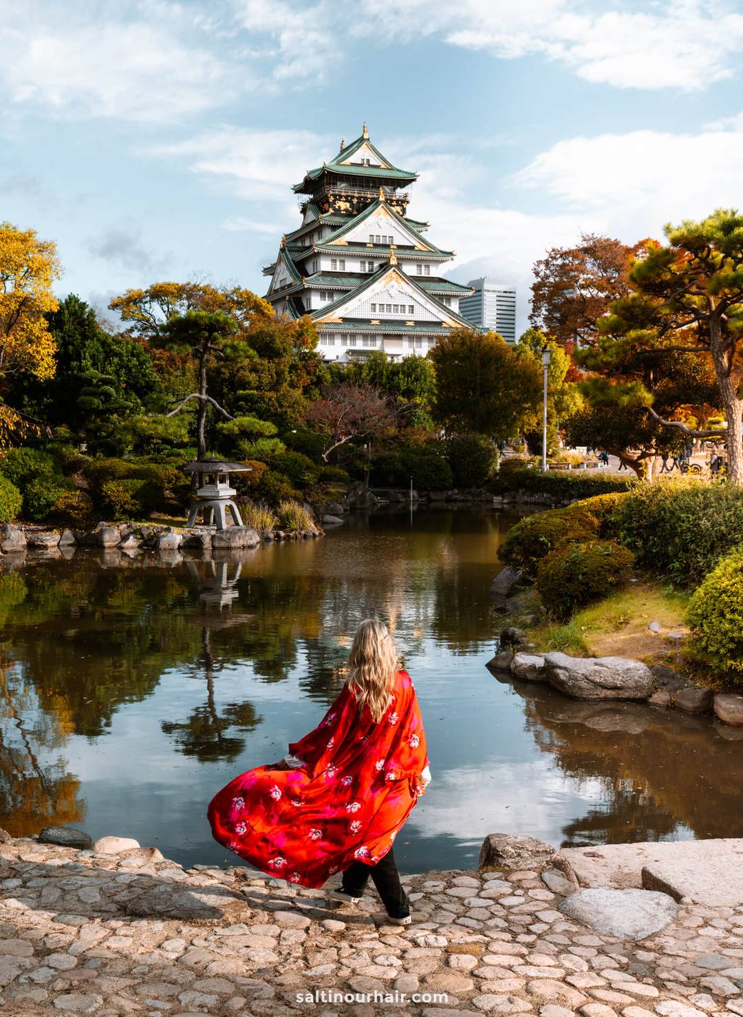 A woman in a red kimono stands by a pond, facing Osaka Castle surrounded by trees and gardens, capturing the serene beauty of autumn in Japan.
