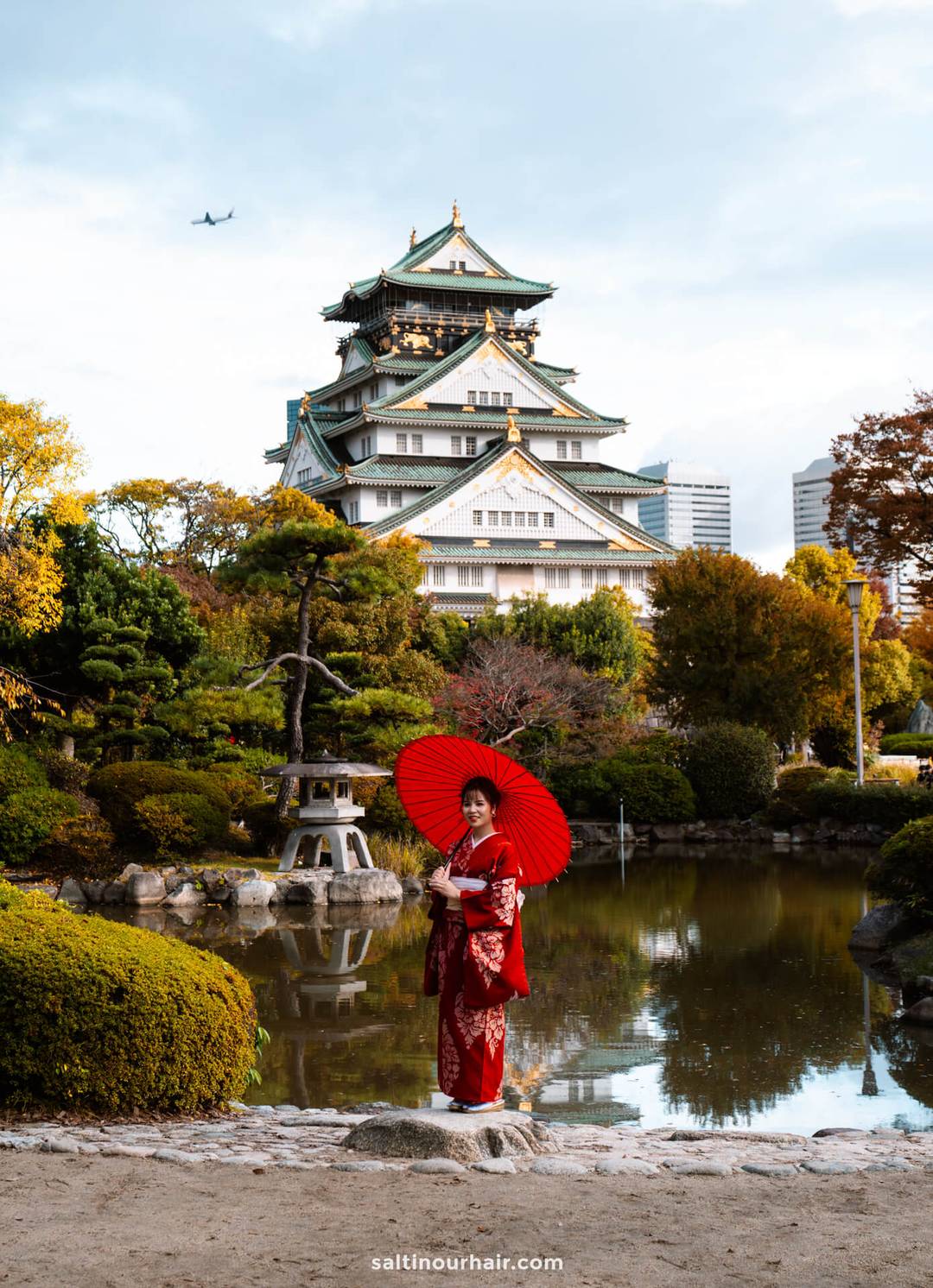 A woman in a red kimono stands by a pond with a red umbrella in Osaka Castle Nishinomaru Garden, surrounded by autumn trees and gardens.