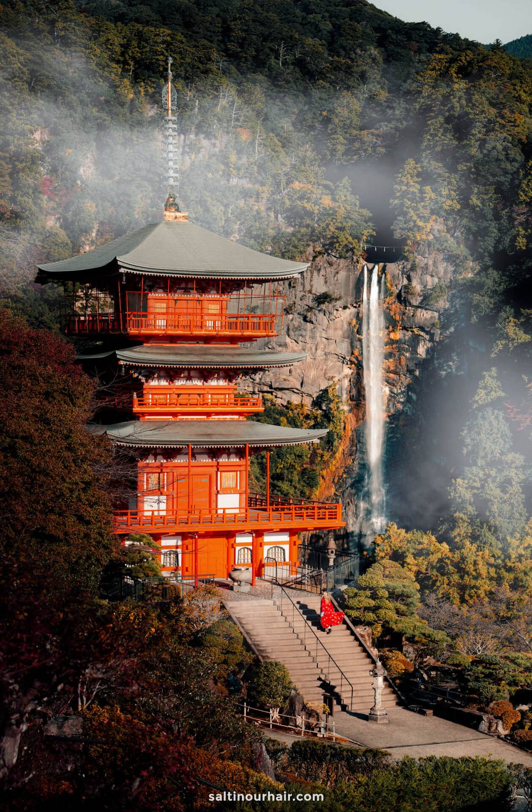 A red pagoda stands among lush greenery with Nachi Waterfall, Japan&rsquo;s tallest waterfall, cascading down a rocky cliff in the background.