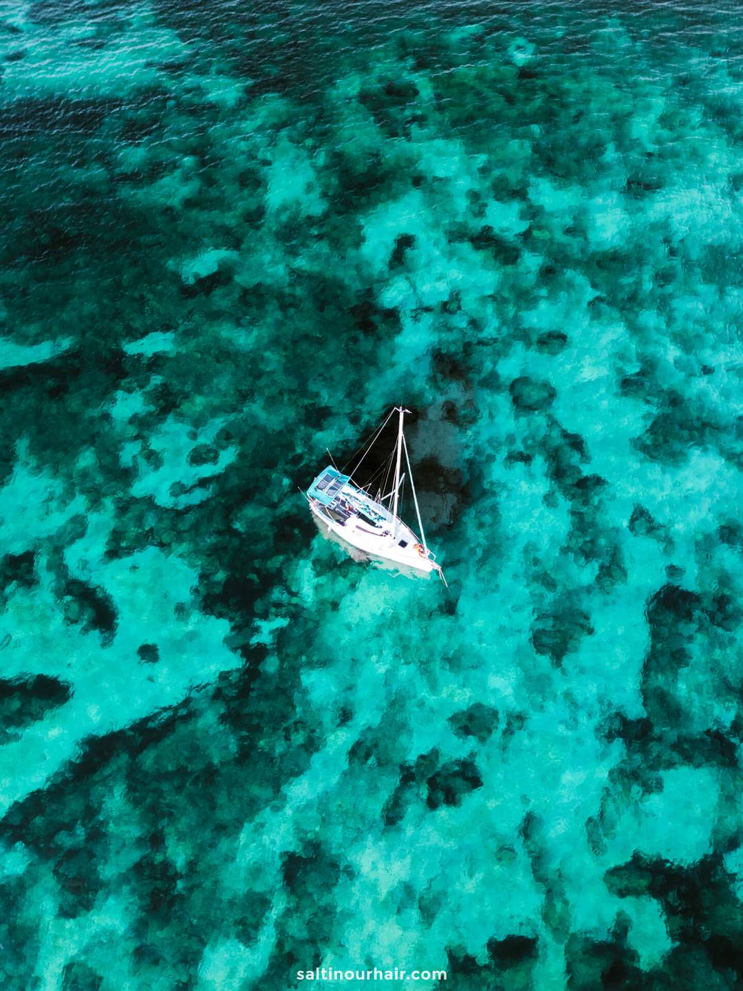 A white sailboat floats on clear, turquoise water with visible dark patches, viewed from above—perfect for island hopping Azores adventures.