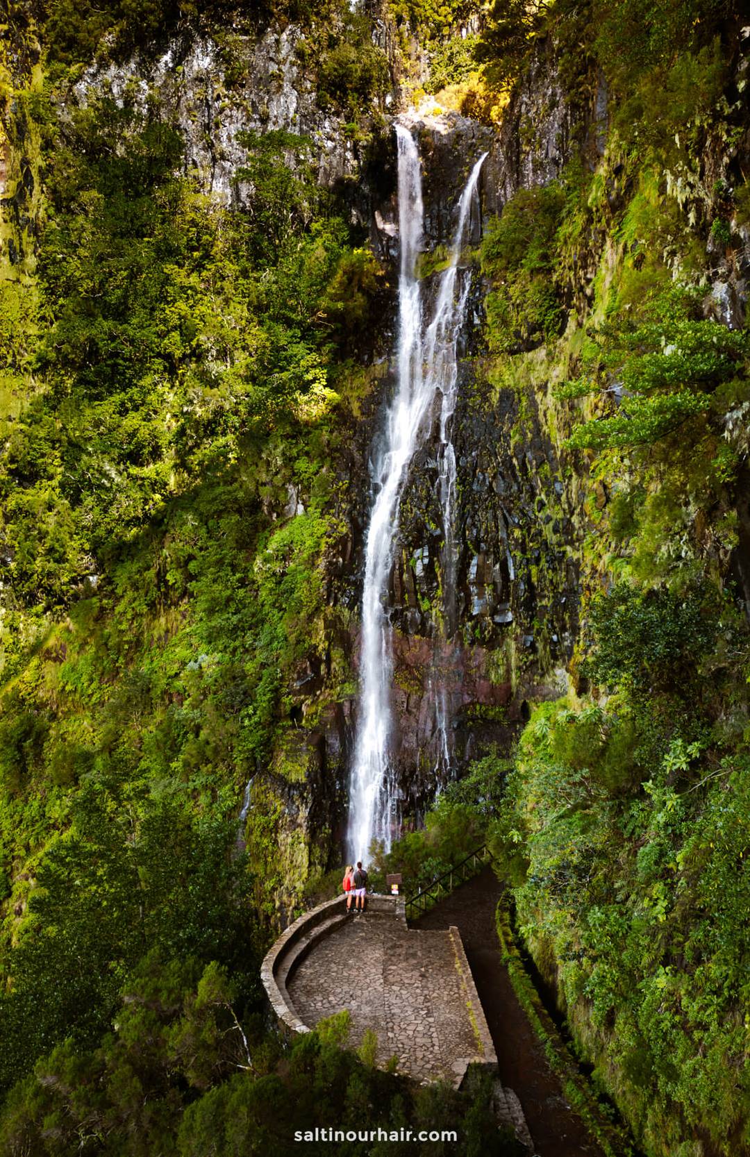 Een hoge, smalle waterval klettert naar beneden langs een weelderige groene klif en eindigt bij een stenen uitkijkplatform waar twee mensen staan &mdash; perfect voor reizigers die benieuwd zijn naar de wandelkosten op Madeira.